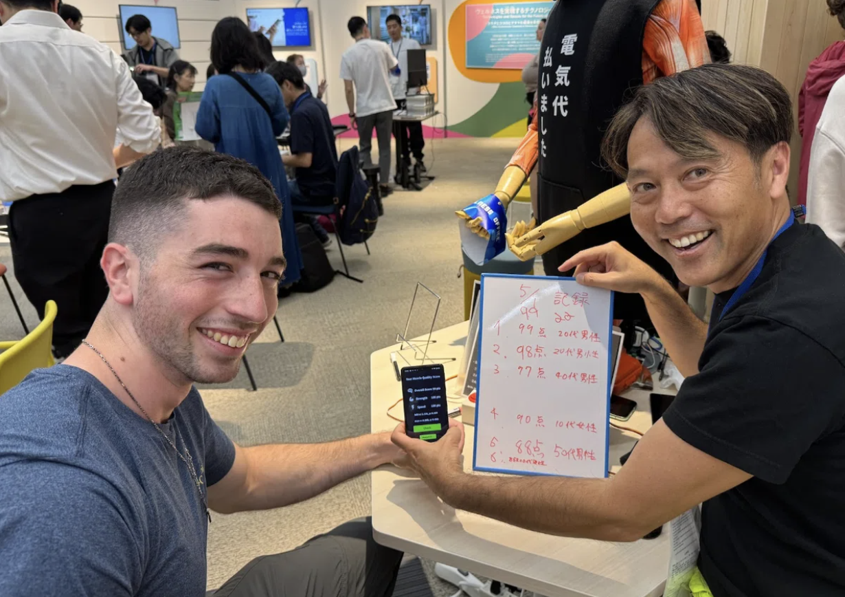 大阪・関西万博の会場で筋肉の質スコアを示すスマートフォンと記録ボードを持つ衣笠竜太さんたち